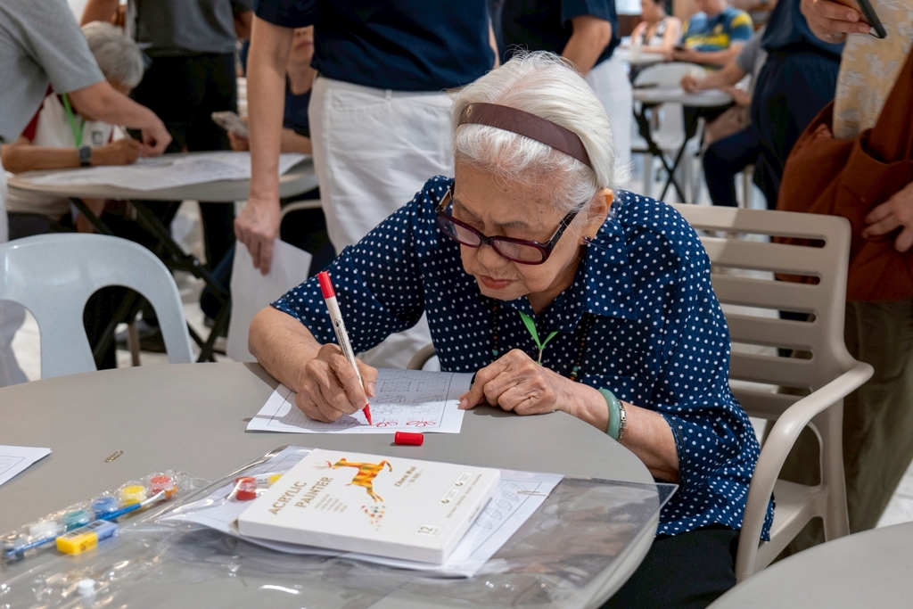A 90 years old senior traces patterns on a worksheet with a colored pen. The goal is to exercise the fine motor skills, or the use of small muscles in the hands and fingers.