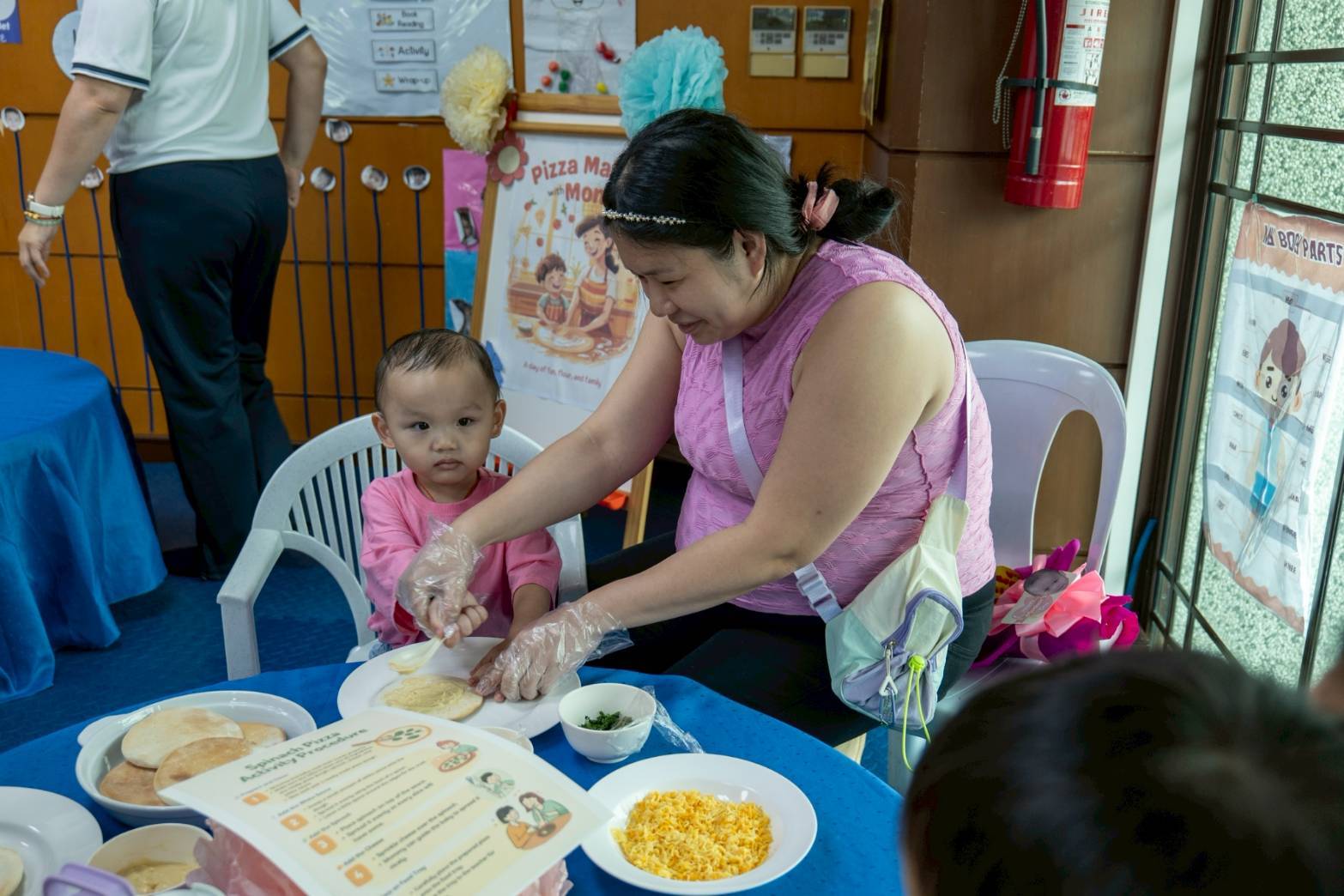 A proud mom carefully guides  her child as they spread cream on pizza dough  together. 