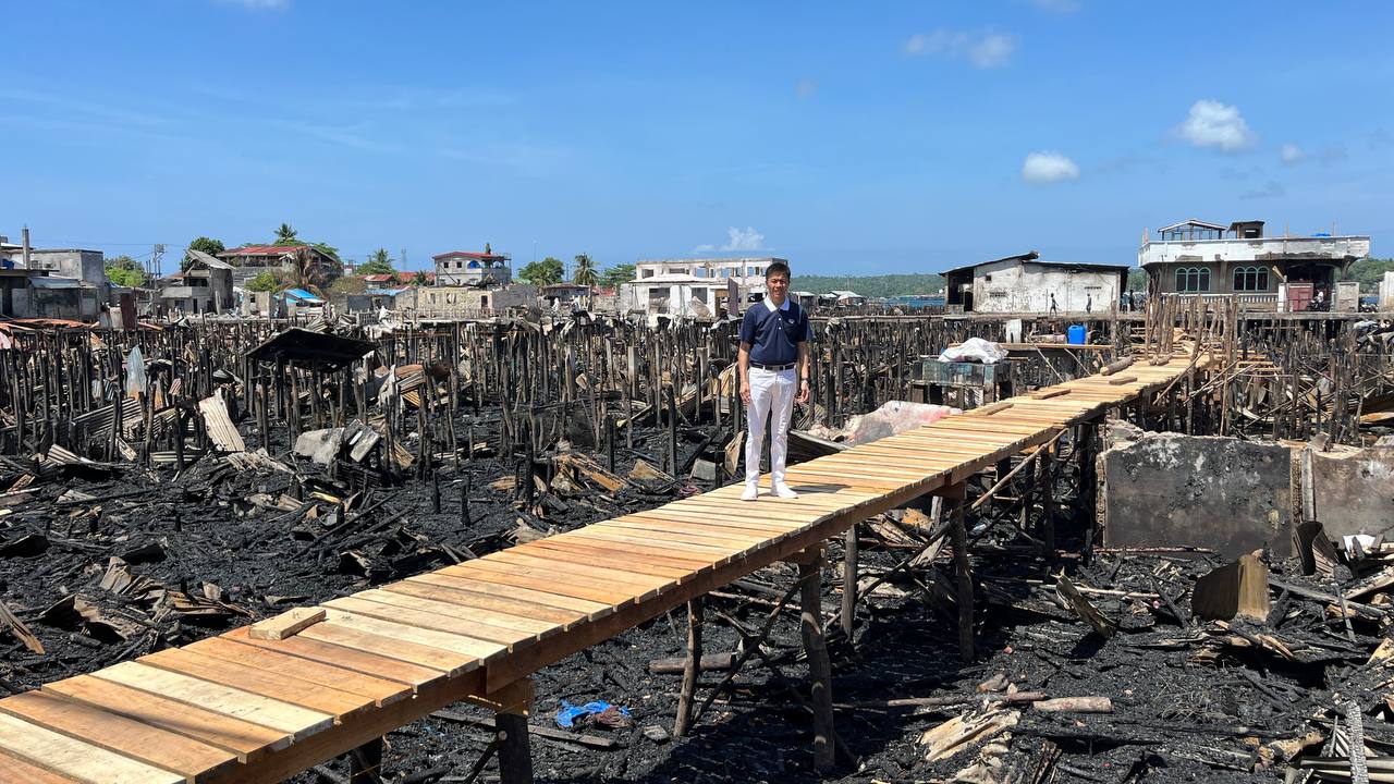 Tzu Chi volunteer Harvey Yap stands along a newly constructed wooden pathway while debris from houses destroyed by the fire can be seen behind him. 