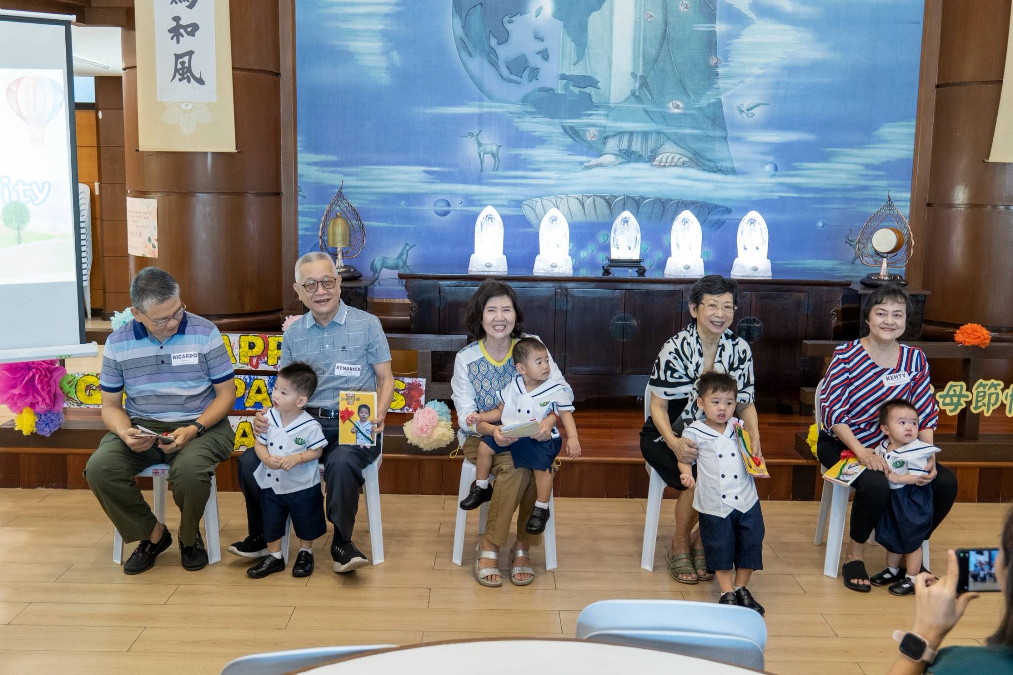 In a heartwarming moment, on stage, the young toddlers happily hand over handmade cards to their beloved grandparents.