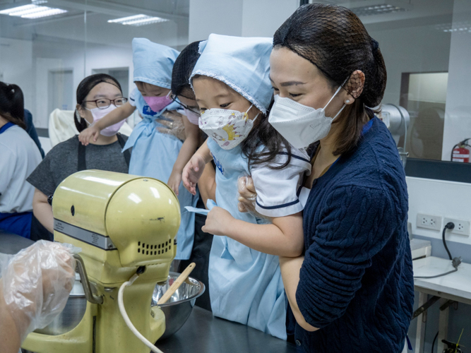 A parent carries her daughter to witness the beating of the mixed ingredients for optimal volume and texture while being cautious of her child’s safety from the electric mixer.