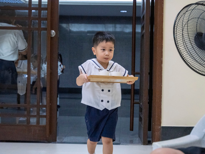 A preschool student enthusiastically looks for his parents while carrying the tea he will offer to them.
