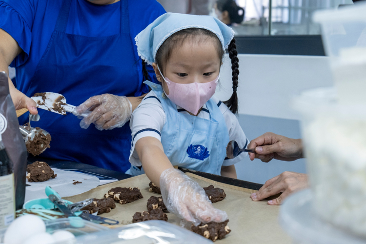 Harmony in baking: As the parent scoops out the cookie dough from the kitchen scale, guaranteeing a 50-gram net weight, this student presses the ball-sized dough on a baking sheet.