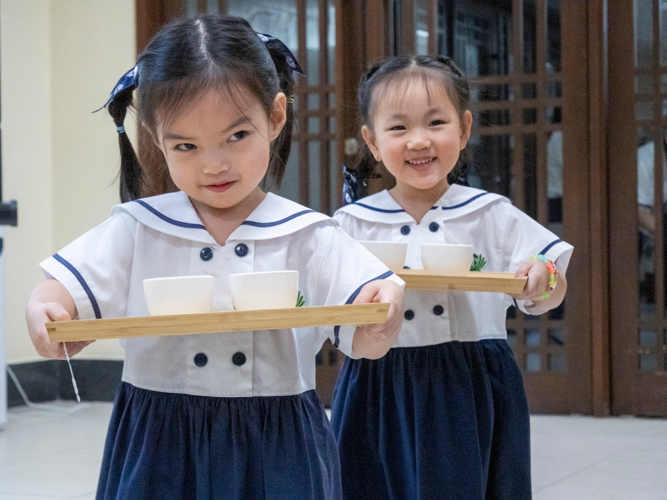Two preschool students happily smile while carrying their trays with care.