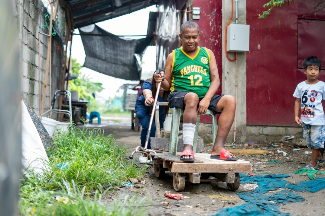 As Levy Telecio's condition makes walking increasingly difficult, his wife, Venus, assists him with a Monobloc chair fixed atop a makeshift wooden trolley. Holding on to his cane for balance, Levy carefully settles into the seat as his wife guides him along.